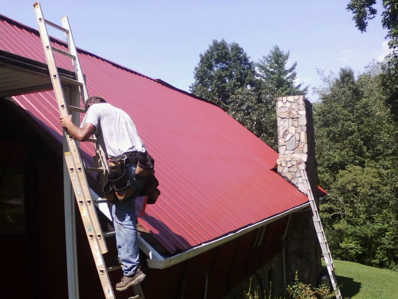 Roofer on a ladder beside a red metal roof and stone chimney — Soffit & Fascia Repair by Roofing Brothers in Pine Island